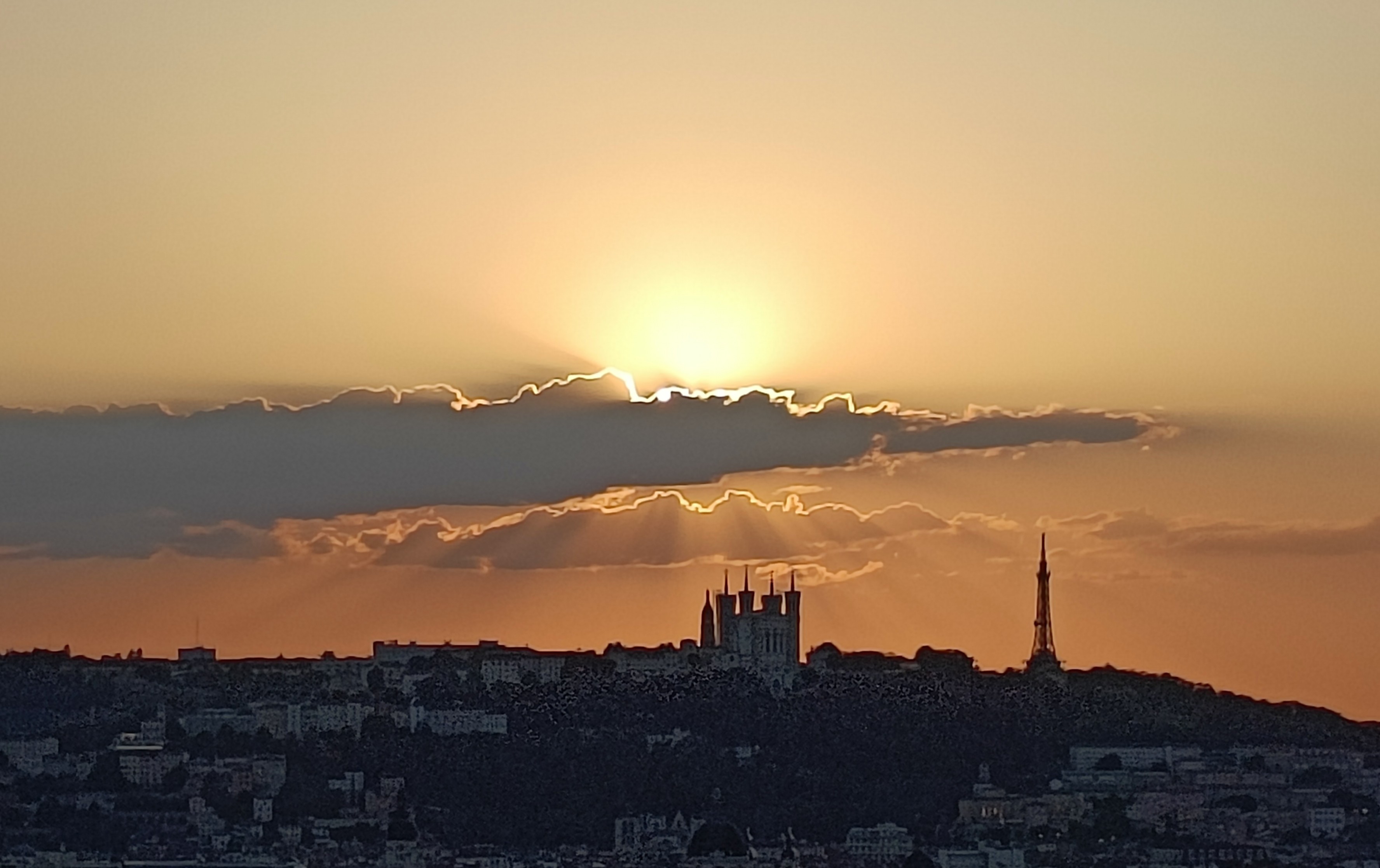 Photo couleur en format paysage du ciel avec la lumière du soleil, atténuée par l'éclipse partielle, qui éclaire le bord de 2 nuages allongés et illumine le ciel en haut du nuage le plus foncé. On aperçoit la silhouette d'une colline avec la basilique de Fourvière et la tour métallique.