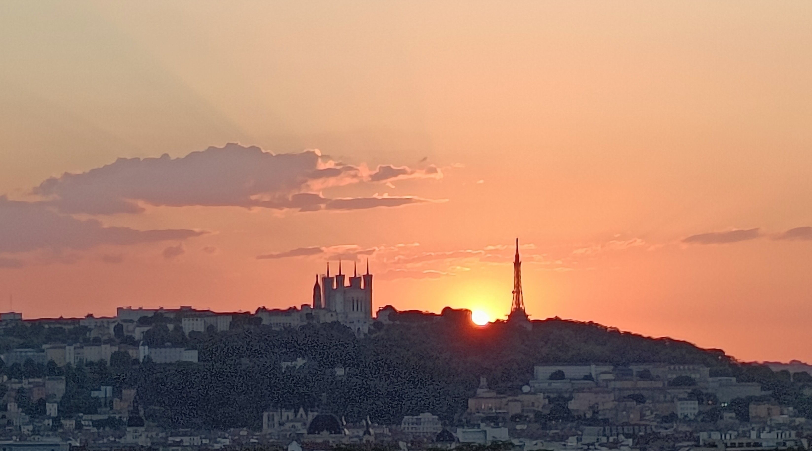 Même type de vue mais au soleil couchant qui disparaît à gauche de la tour métallique. Le ciel est rose orangé avec quelques nuages.