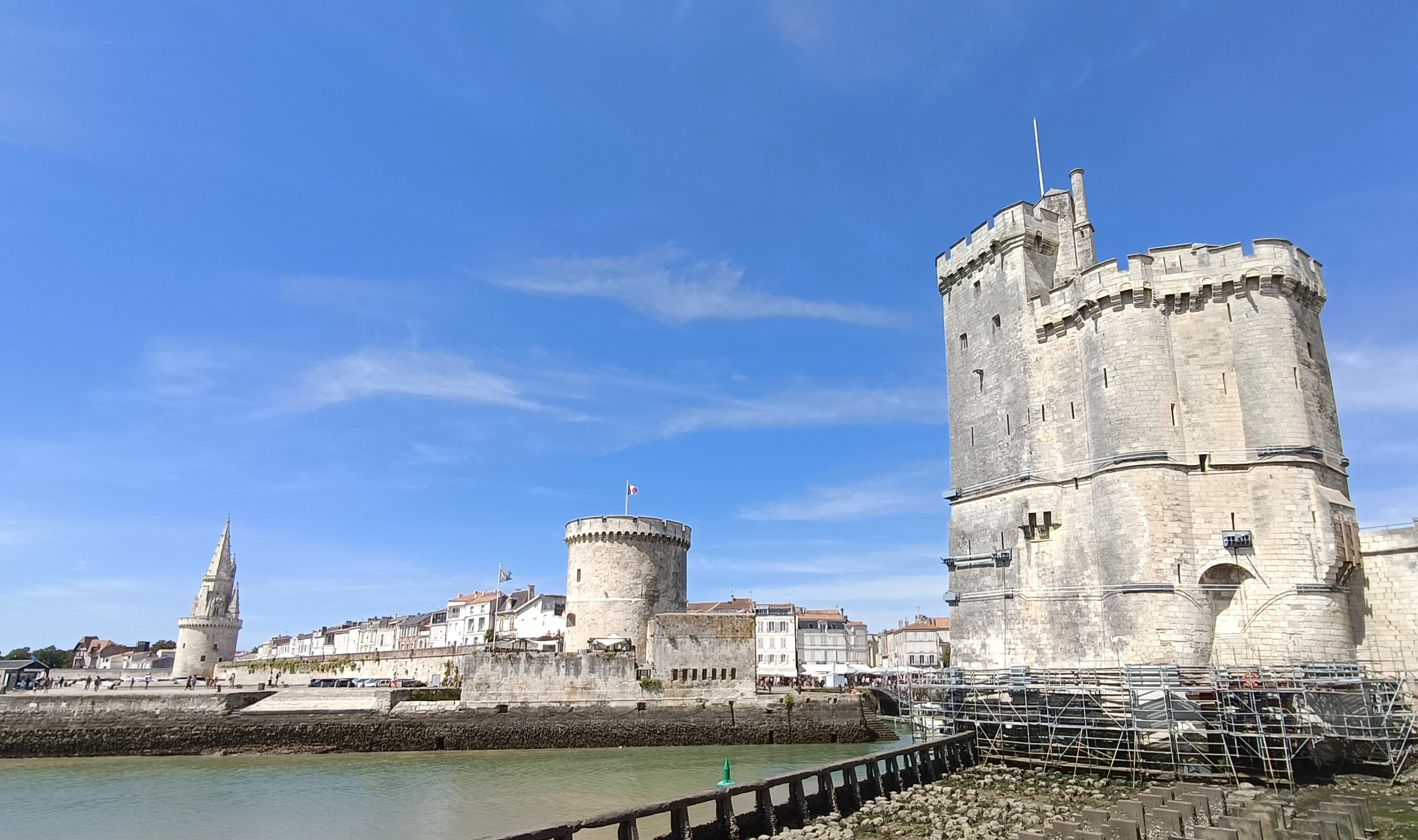 4 photos couleur de différents formats. Vue panoramique de l'entrée du port de la Rochelle, avec 1 tour de chaque côté qui garde l'entrée. La tour Saint-Nicolas à droite penche et est ceinturée de bandages en fer. A gauche, une autre tour avec un clocher pointu, le tout en pierres de taille claires. Devant, l'océan à marée basse et une balustrade en fer. Le ciel est bleu, lumineux avec quelques nuages d'altitude.