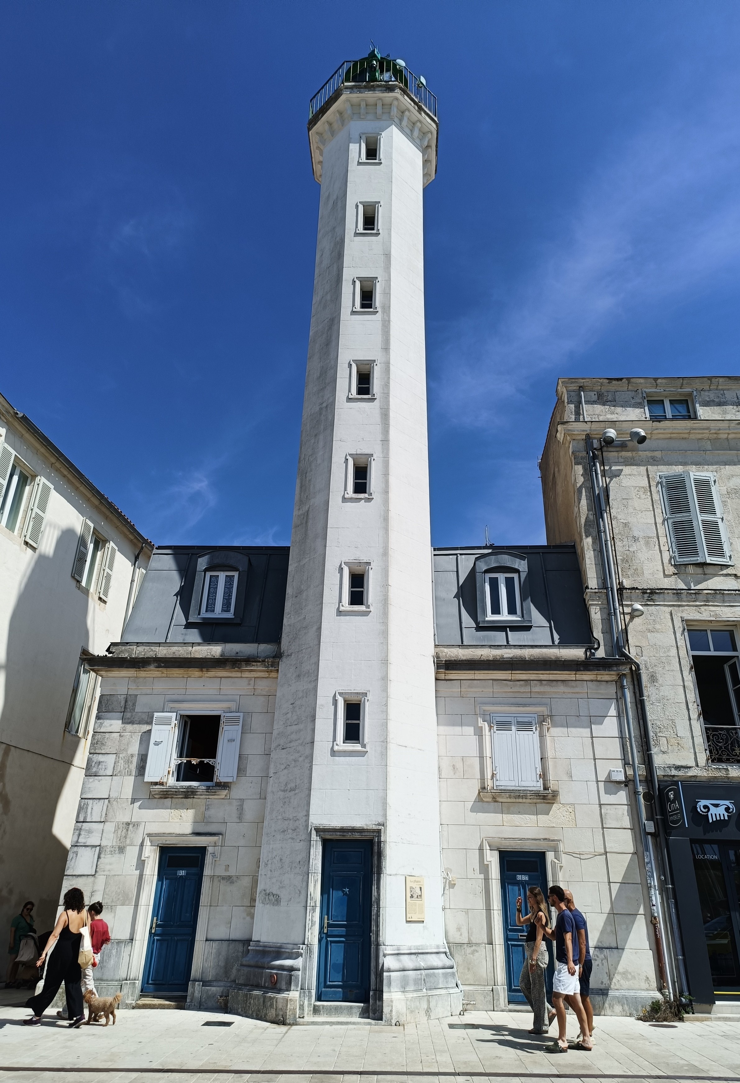 Vue en contre-plongée d'un grand phare blanc au sommet vert, encastré dans une maison de 2 étages au toit en zinc et en pierres calcaires. Des immeubles un peu plus haut de part et d'autre et des passants devant. Le ciel est bleu avec des nuages d'altitude.