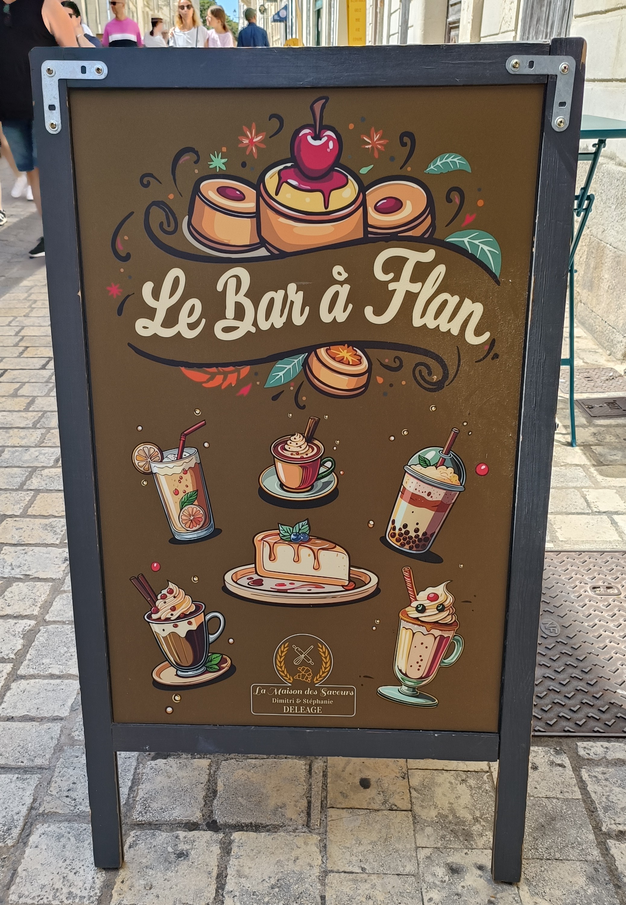 Dans une rue pavée en pierres calcaires, un présentoir à 2 pans présente "Le bar à flan". Sur un fond marron sont dessinés des gâteaux et des boissons en couleurs vives.