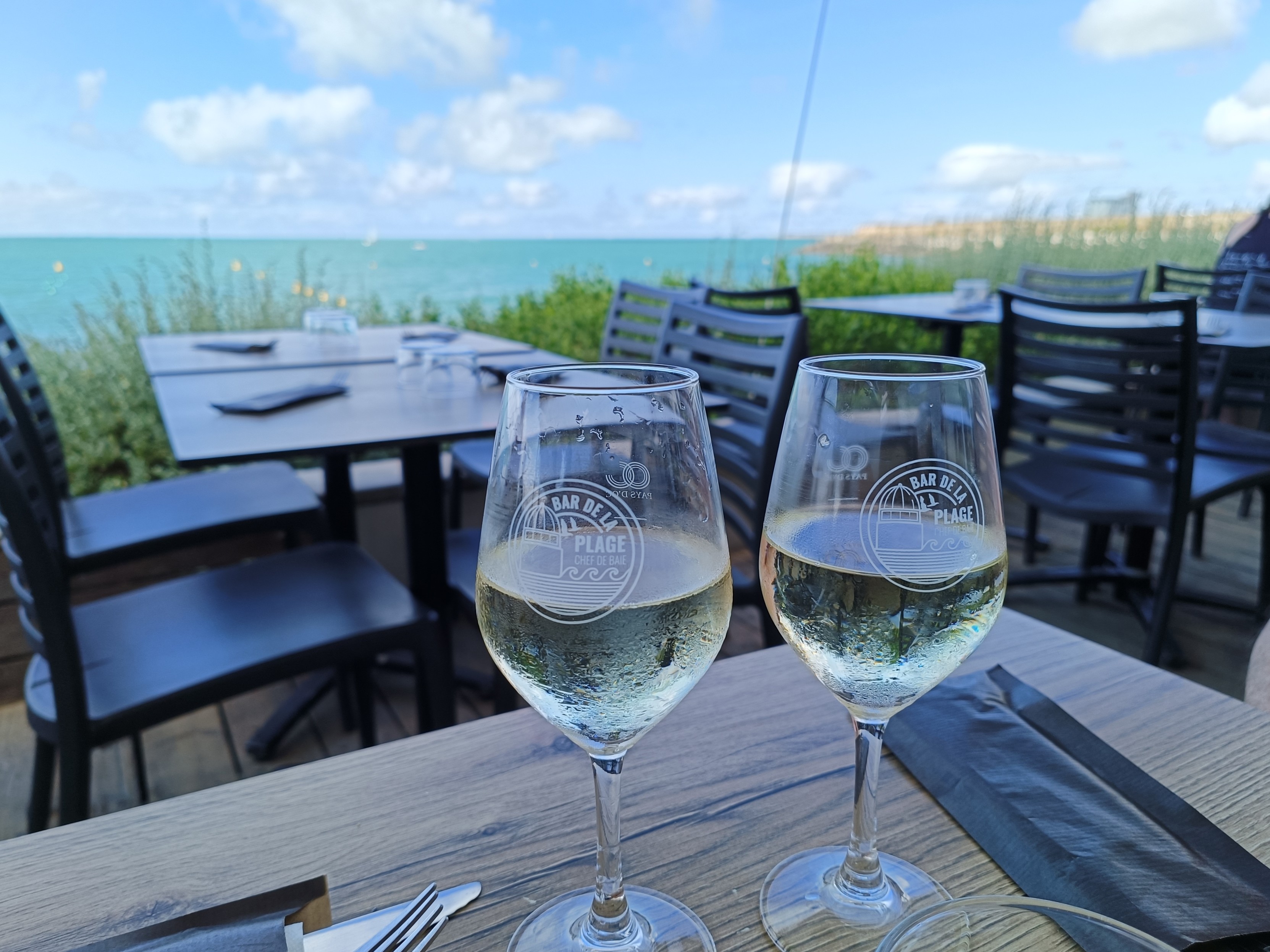 Vue rapprochée de 2 verres de vin blanc posés sur une table claire, sur laquelle sont posés des couverts. On voit d'autres tables vides et l'océan au loin. Le ciel est bleu clair avec des nuages blancs.