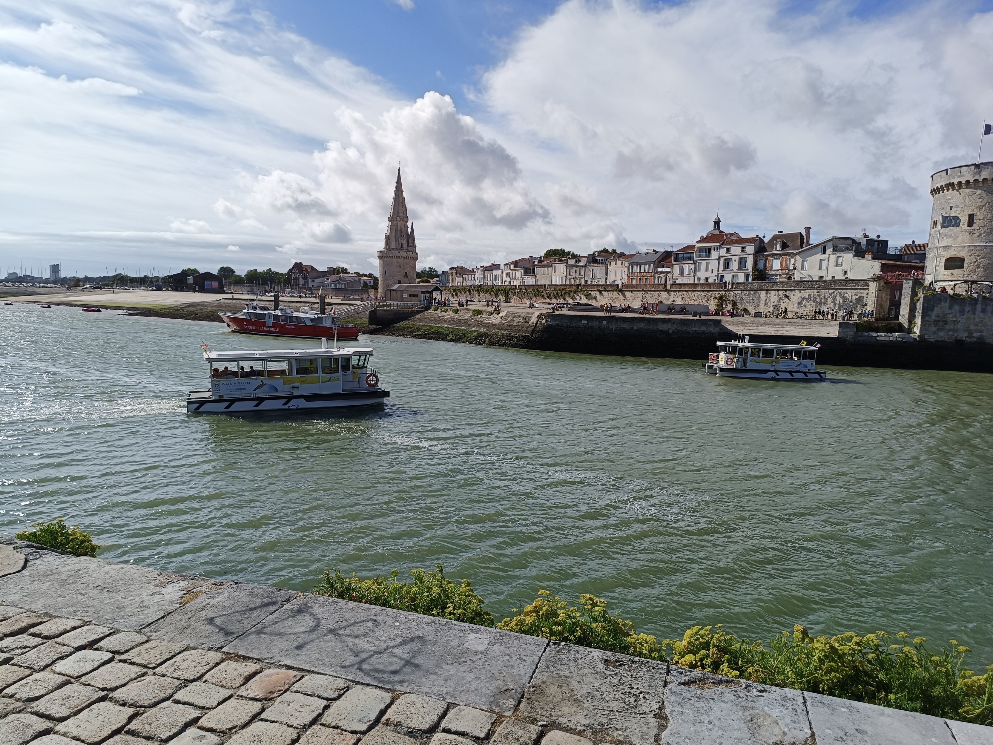 Vue prise depuis un quai en pavés, avec sur l'eau 2 navettes fluviales blanches qui vont se croiser. Sur la rive en face, on aperçoit une muraille, une tour ronde surmontée d'un clocher gothique (la tour de la lanterne) et une tour ronde crénelée (la tour des chaînes). Le ciel est bleu, lumineux avec des nuages blancs.