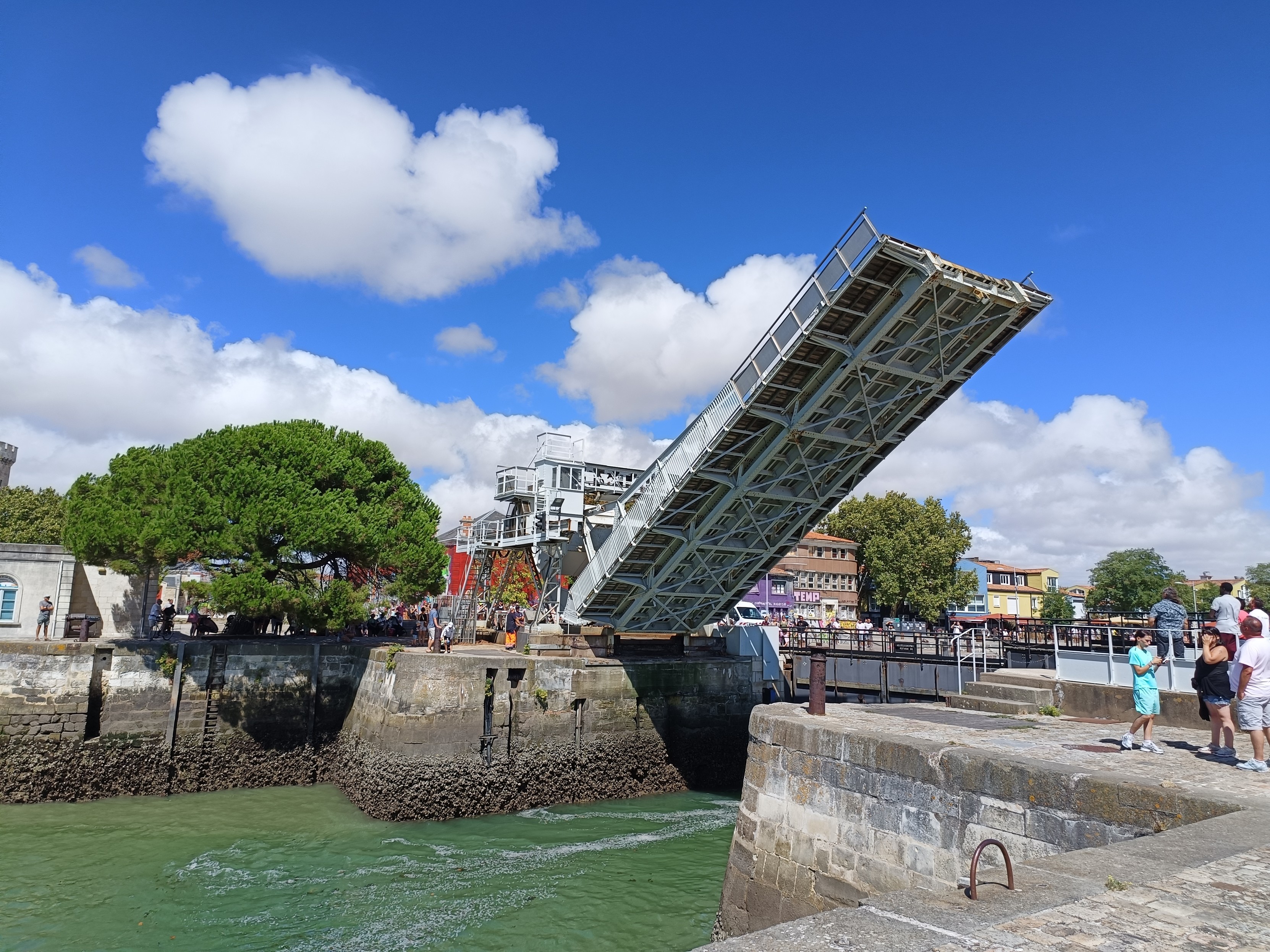 Vue d'un pont levant (du Gabut) dont le tablier est relevé à 45 degrés environ. Il enjambe l'entrée d'un des bassins, qui est fermé par une écluse. On voit des gens qui observent et attendent pour traverser, un pin maritime et le ciel bleu avec des nuages blancs.
