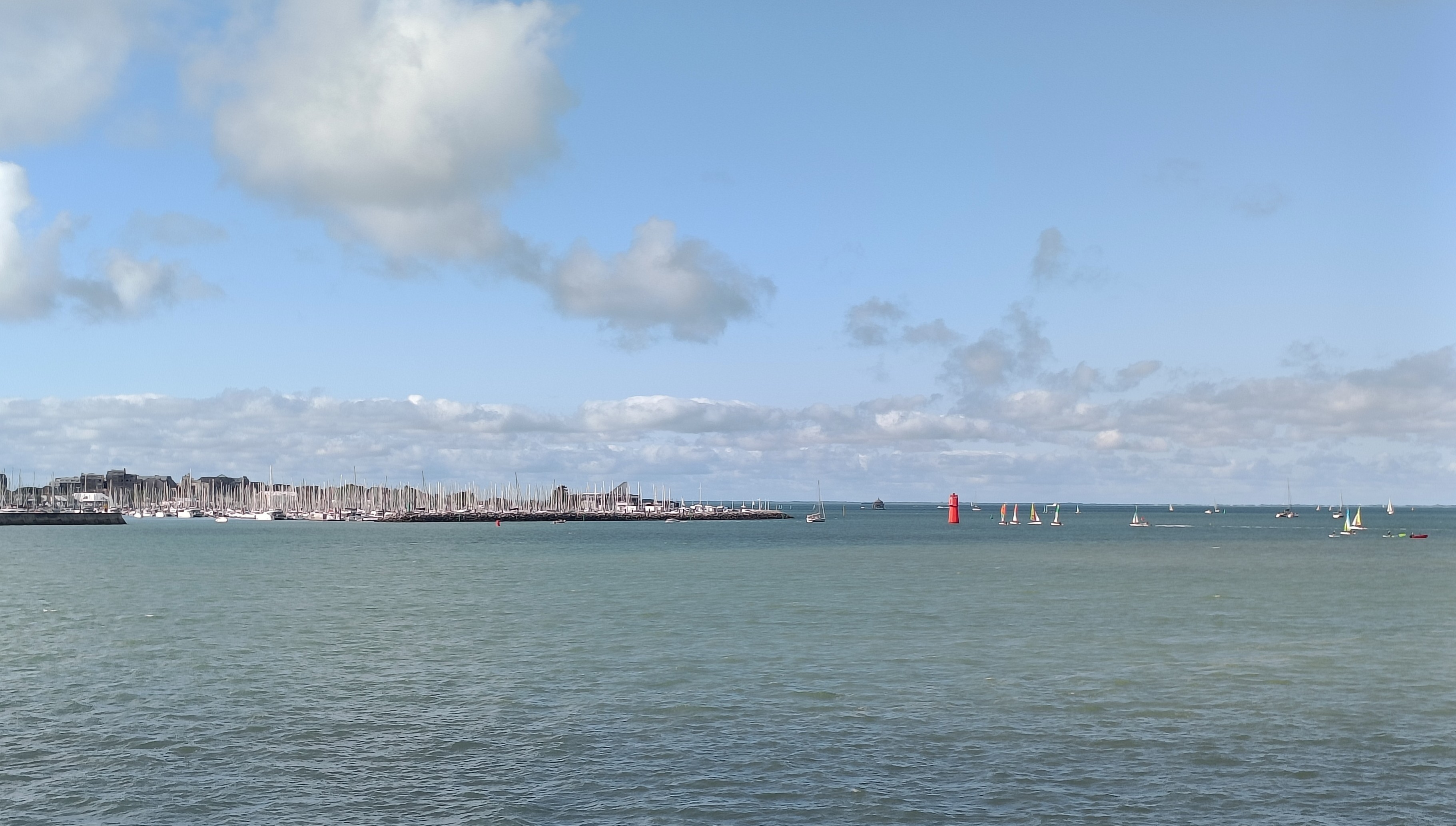 4 photos couleur de différents formats. Vue panoramique de l'océan en direction de la plage des Minimes, où l'on aperçoit de nombreux mâts de bateaux. Sur l'eau, une bouée rouge et des voiliers de petites tailles, dont des bateaux écoles. L'eau est bleu-vert, le ciel est bleu clair avec des nuages blancs et gris, dont certains forment une bande sur l'horizon.