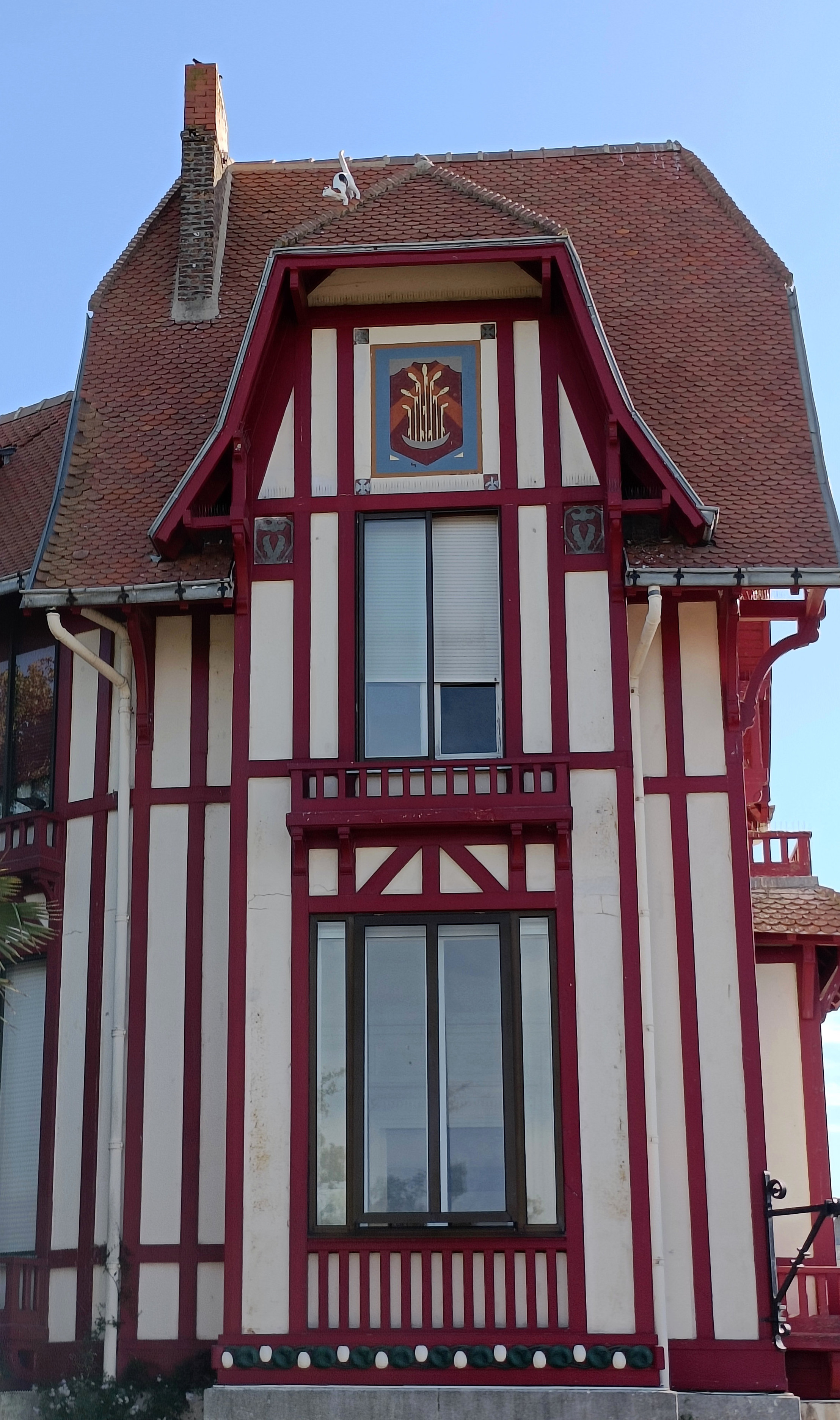 Photo couleur en format portrait, avec la vue en légère contreplongée de la façade d"une villa de 2 étages avec des pans de bois rouges et des murs clairs, 2 fenêtres, un décor d'épi sur fond bleu au-dessus de la fenêtre supérieure. Sur le toit très pentu et au sommet d'une avancée, une sculpture de chat noir et blanc. Il a le dos arrondi et la queue levée, on voit son profil gauche. Le ciel est bleu.
