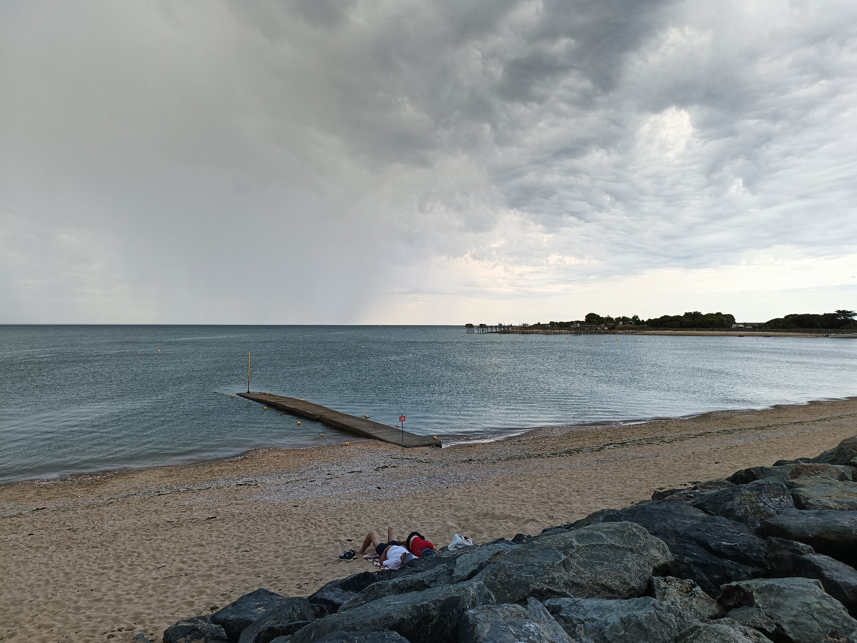 Photo couleur en format paysage, avec une vue panoramique prise d'un chemin au-dessus de la plage bordée de gros cailloux noirs. En contrebas, 2 personnes sont allongées sur la plage près des cailloux. Un ponton en bois s'enfonce dans l'océan, une bande de terre à droite délimite l'extrêmité de la baie. 
Le ciel est gris avec des nuages menaçants, on aperçoit un rideau de pluie au loin.
