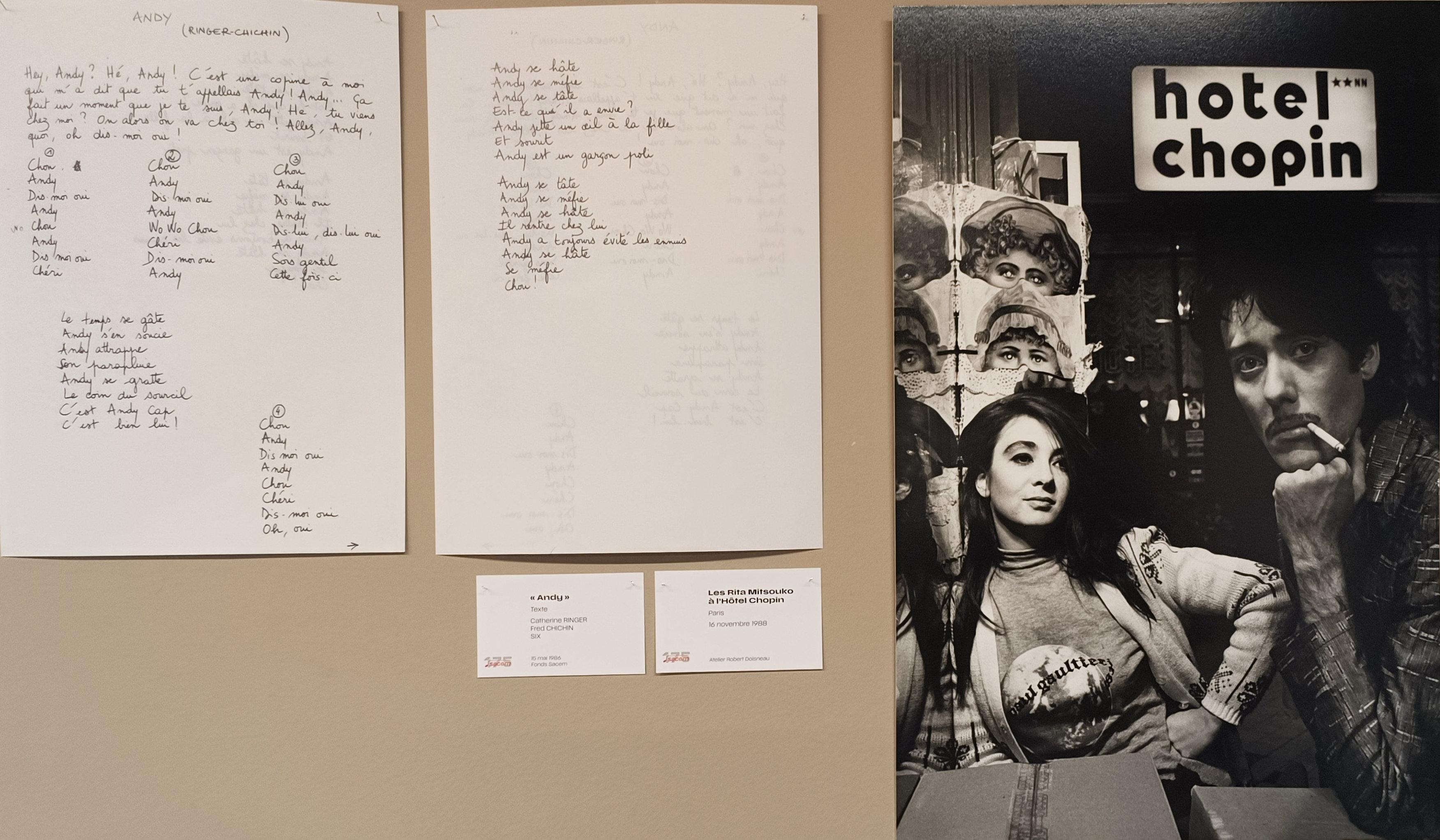 Photo couleur en format paysage, avec sur un mur beige 2 pages blanches écrites à l'encre noire qui sont le texte original de la chanson Andy en 1986. A droite, un tirage photo noir et blanc représente Catherine Ringer avec la main droite sur la hanche et Fred Chichin la clope au bec, avec l'enseigne de l'hôtel Chopin à Paris en 1988. 2 cartels sont accrochés en dessous de la 2e feuille blanche.